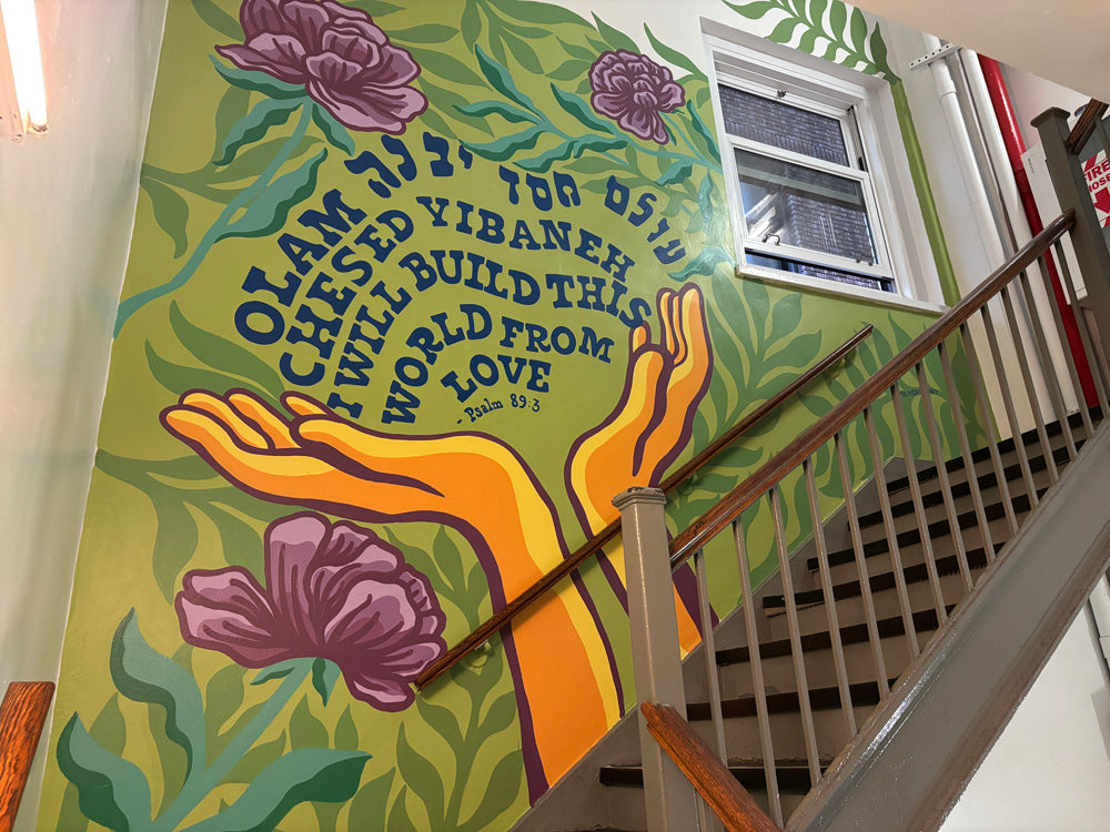 A colorful mural in a stairwell that features two upraised hands cupping a quote that says "olam chesed yibaneh I will build this world from love" and large purple flowers