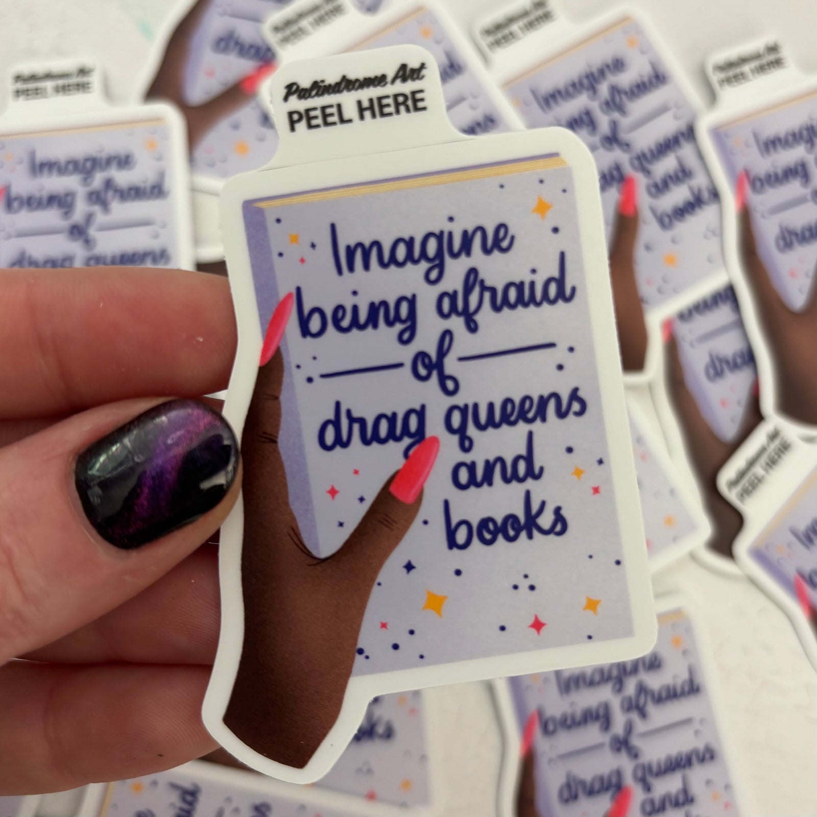Hand holding a sticker with text 'Imagine being afraid of drag queens and books' among more stickers.