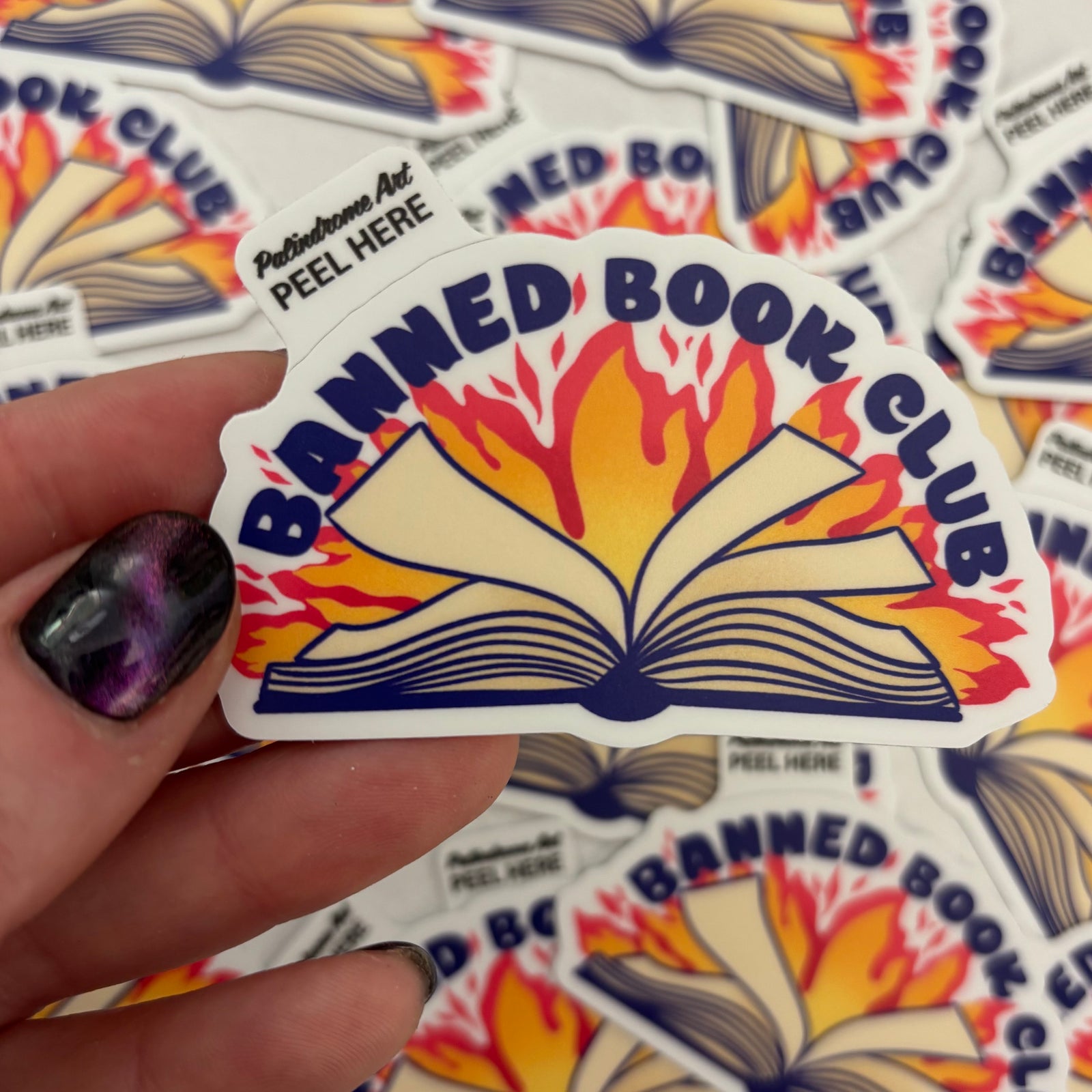 Sticker with 'Banned Book Club' design held by a hand, with more stickers in the background.