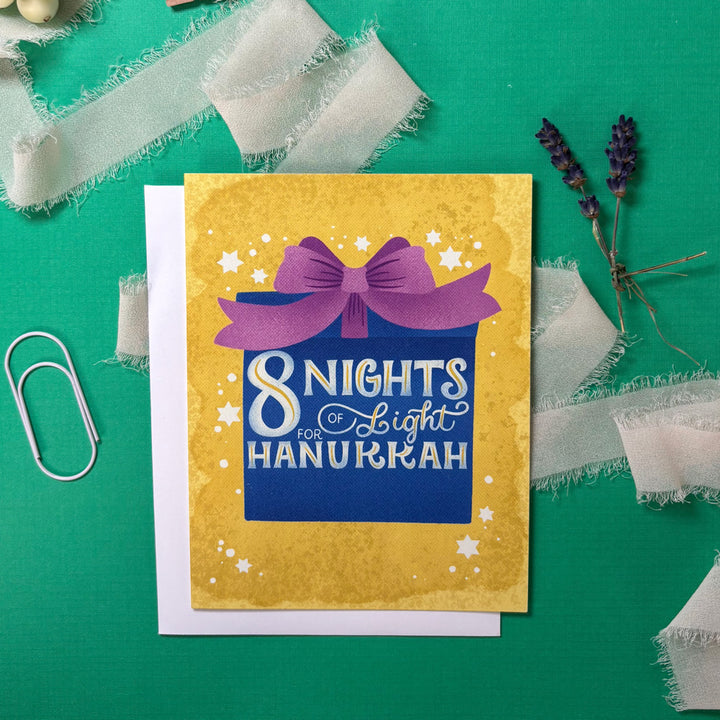 Hanukkah card with purple bow on yellow background, surrounded by decorative elements on a green surface.
