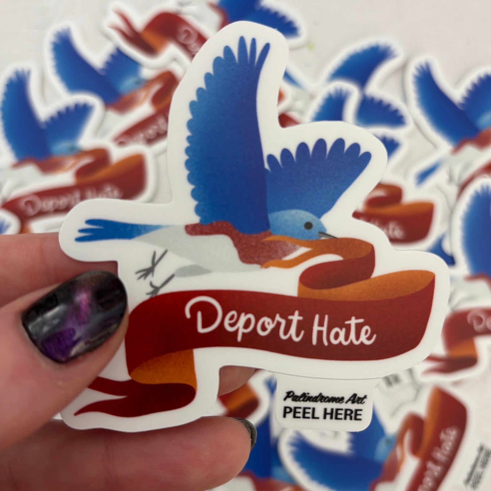 Sticker with a bird and 'Deport Hate' text held by a hand, with more stickers in the background.