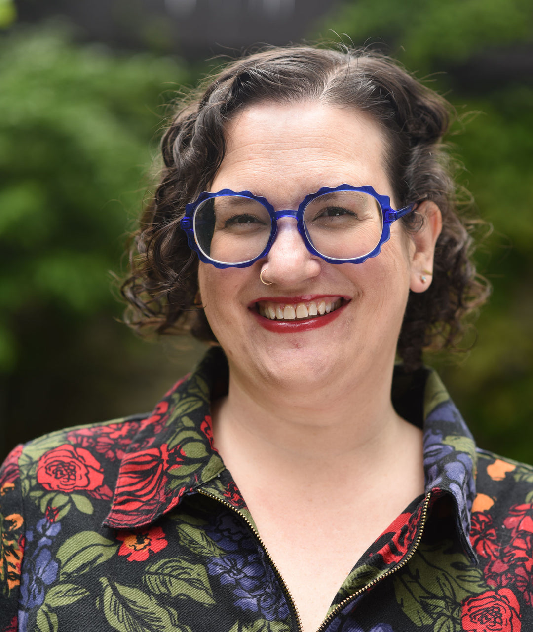 a woman with short curly hair and blue glasses, with a nose ring, wearing a flower jumpsuit