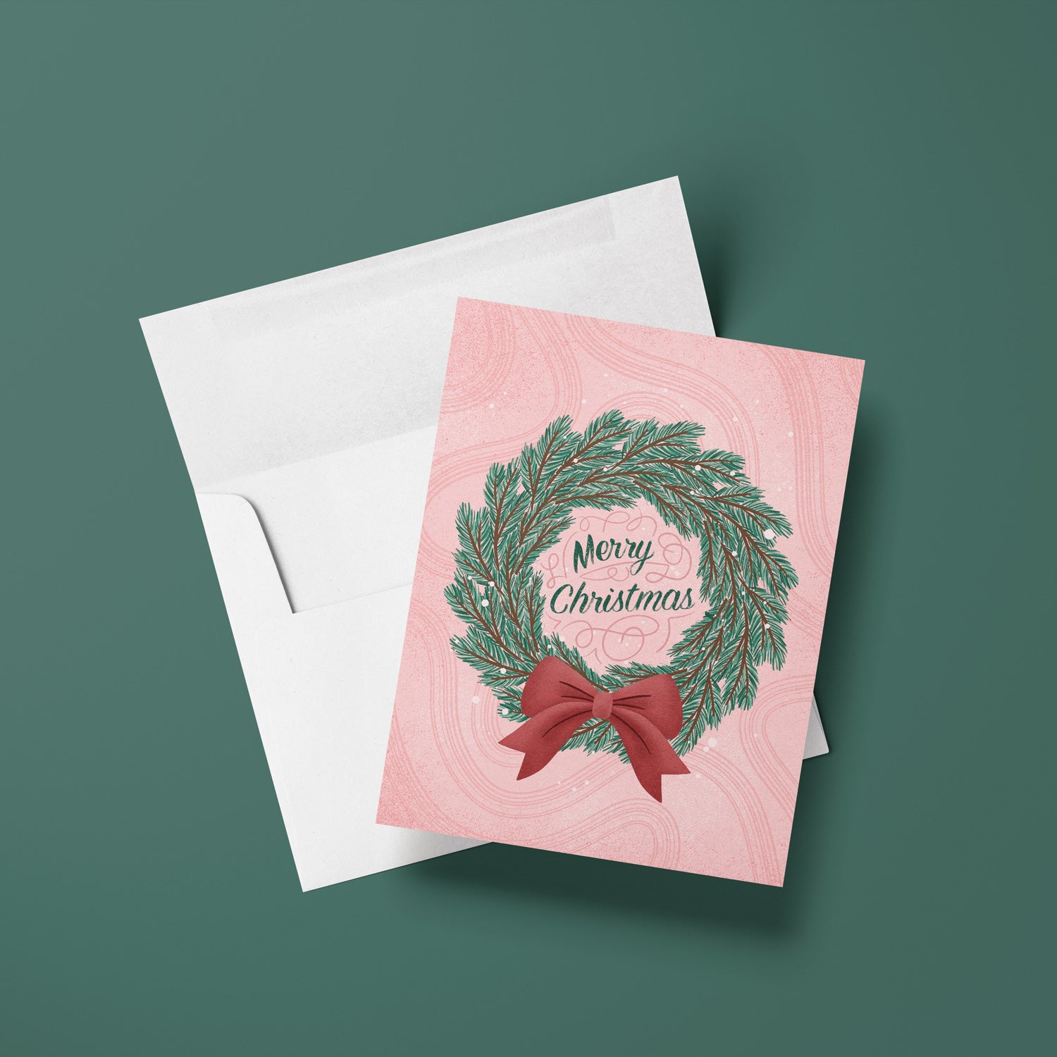 Pink Christmas card with wreath and bow design on a white envelope against a green background
