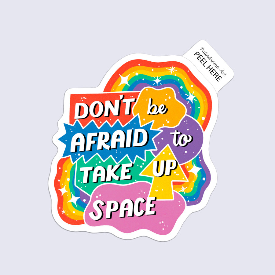 A colorful die-cut sticker with the phrase “DON’T be AFRAID to TAKE UP SPACE” in bold white lettering over rainbow and starburst shapes, shown on a light gray background. A small tag at the top reads “Palindrome Art Peel Here.