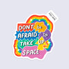 A colorful die-cut sticker with the phrase “DON’T be AFRAID to TAKE UP SPACE” in bold white lettering over rainbow and starburst shapes, shown on a light gray background. A small tag at the top reads “Palindrome Art Peel Here.