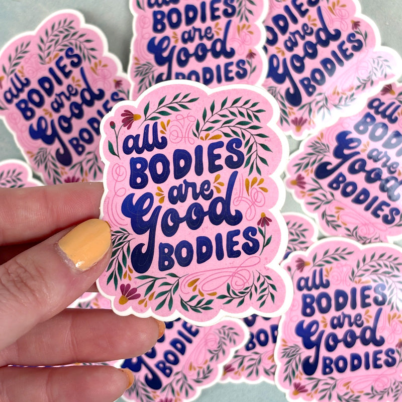 hand holding A sticker with a pink background and a green floral pattern, featuring the text 'All Bodies are Good Bodies' in a bold, cursive font.