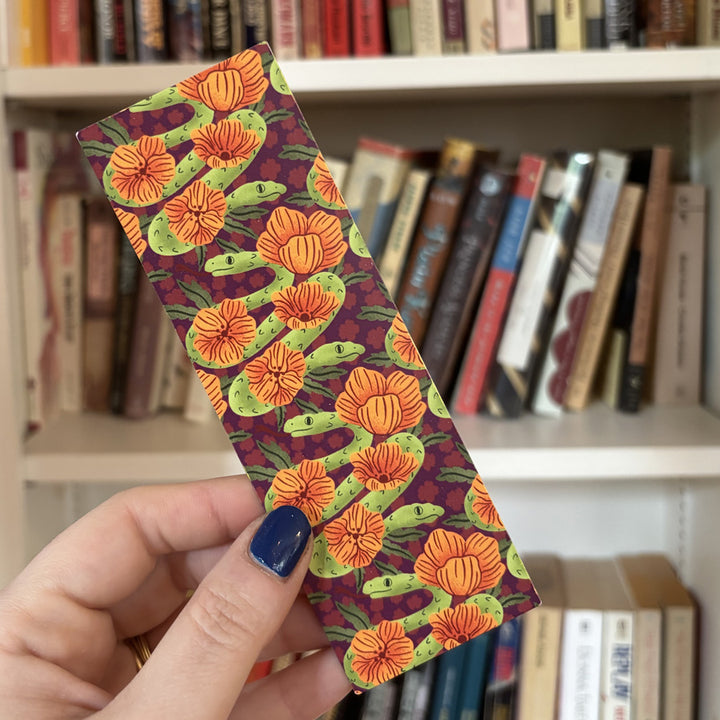 hand holding a bookmark with an illustration of a two headed snake in front of a bookcase