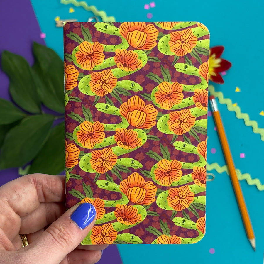 hand with blue nail polish holding a small notebook with rounded corners featuring a pattern of illustrated two headed snakes and orange flowers