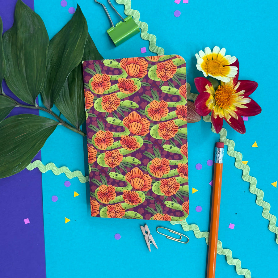a styled flat-lay photo of a small notebook with rounded corners featuring a pattern of illustrated two headed snakes and orange flowers