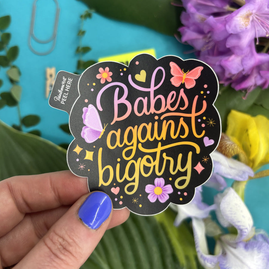 Sticker with 'Babes against bigotry' text held by a hand with blue nail polish, against a floral background.