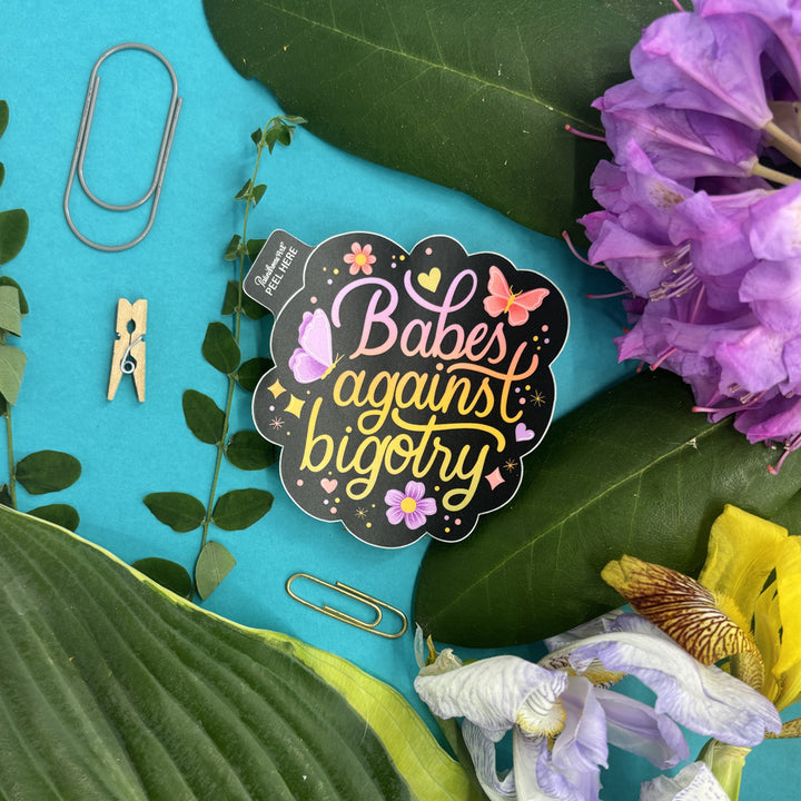 Sticker with 'Babes against bigotry' text on a blue background with flowers and paper clips.