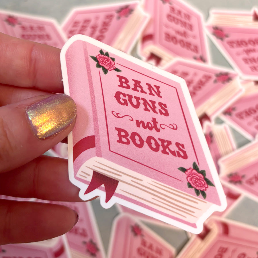 Pink sticker with 'Ban Guns not Books' text held by a hand with glittery nail polish.
