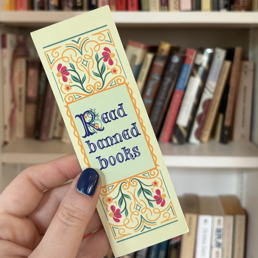 Hand holding a decorative bookmark with 'Read banned books' text in front of a bookshelf.