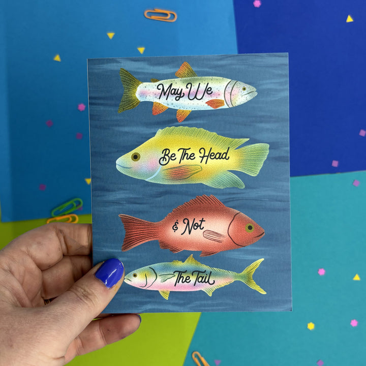 Hand holding a greeting card with illustrated fish swimming in the ocean with text "may we be the head and not the tail"