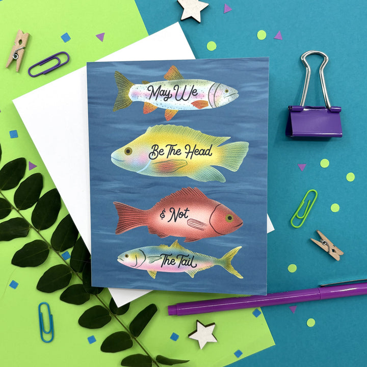 greeting card with illustrated fish swimming in the ocean with text "may we be the head and not the tail" on a colorful background
