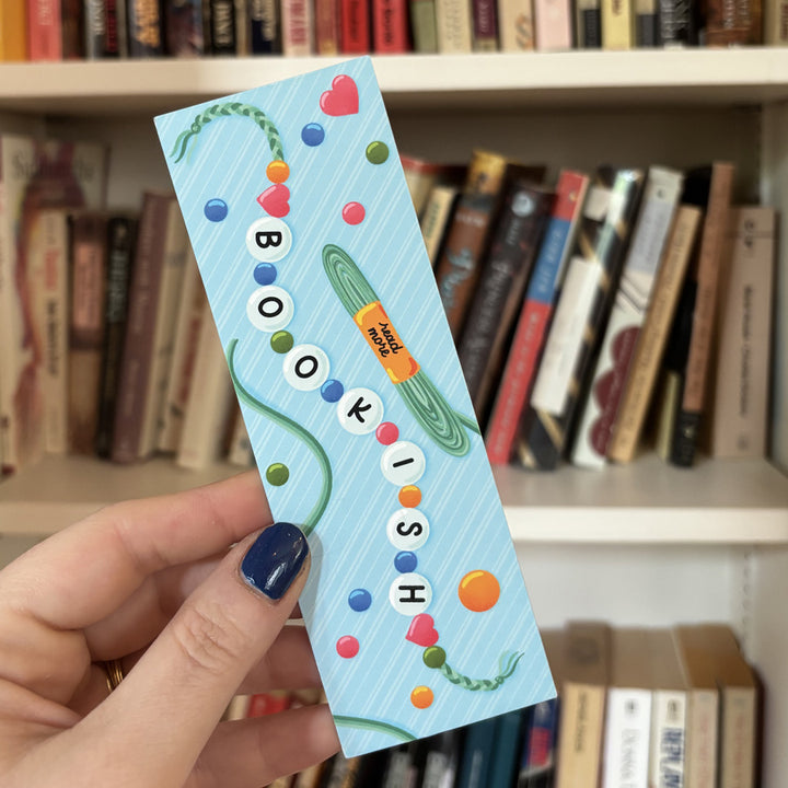 Hand holding a colorful 'Bookish' bookmark in front of a bookshelf.