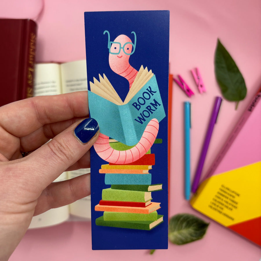 Bookmark with a cartoon worm reading a book, held by a hand on a pink background with stationery items.