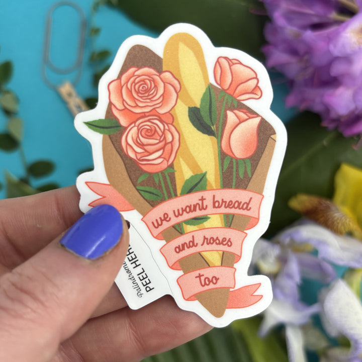 Sticker with bread and roses design and text "we want bread and roses too" held by a hand against a floral background