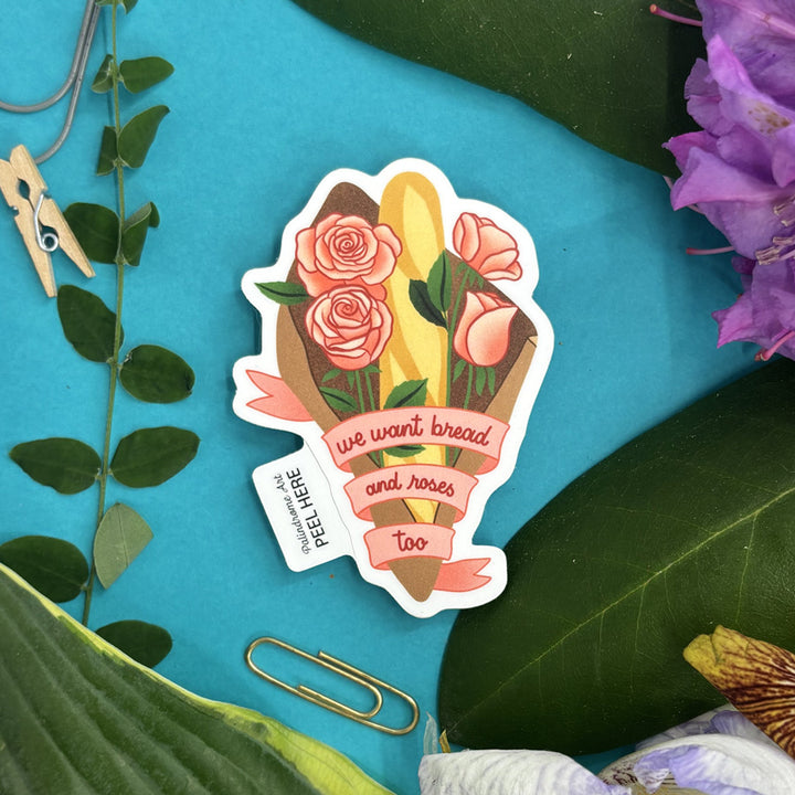 Sticker with bread and roses design and text "we want bread and roses too" on a blue background with leaves and flowers.
