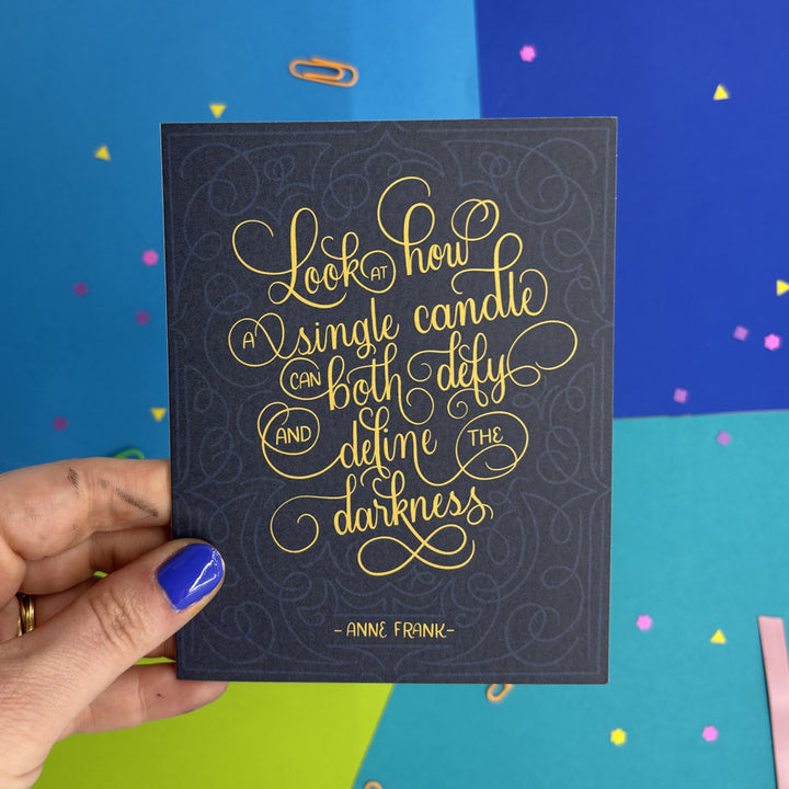 hand holding a hanukkah card with Anne Frank quote "look at how a single candle can both defy and define the darkness"