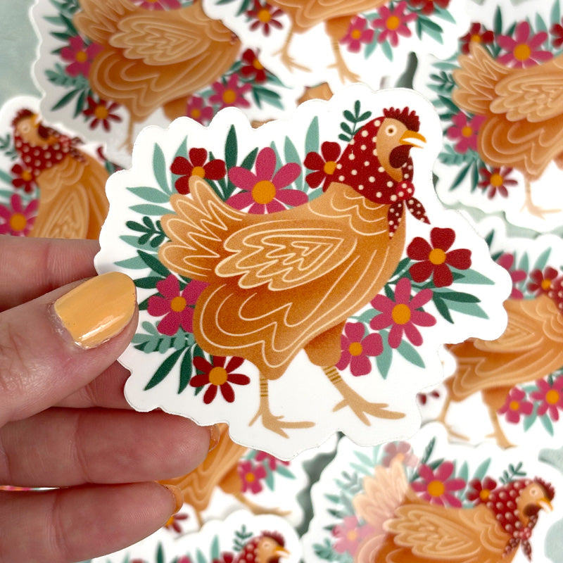 hand holding a sticker of a chicken wearing a polka dot handkerchief in front of flowers