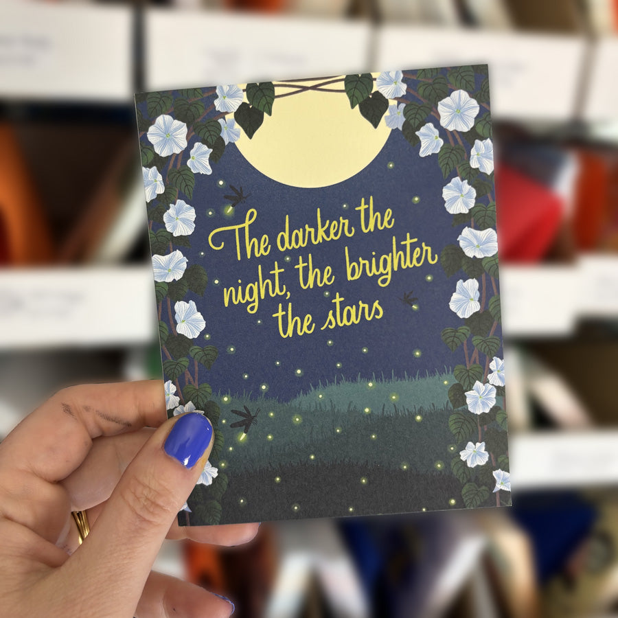 hand holding a greeting card with moonlight flowers at night and text "the darker the night, the brighter the stars"