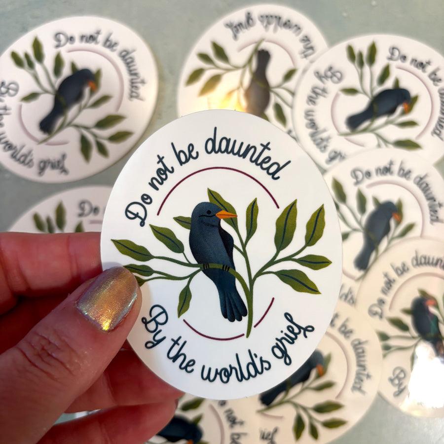 Circular sticker with bird design and text 'Do not be daunted by the world's grief' held by a hand.