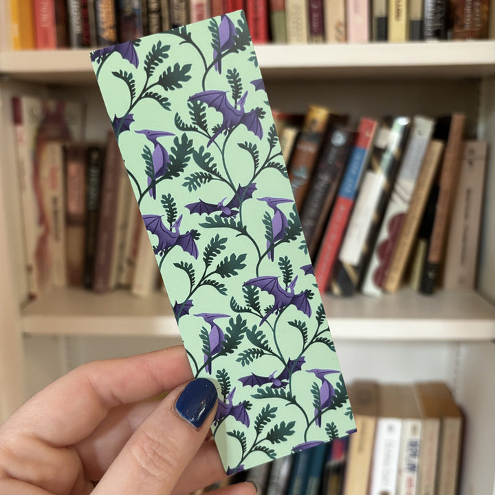 hand holding a bookmark featuring purple pterodactyl dinosaurs and leafy plants in front of a bookshelf