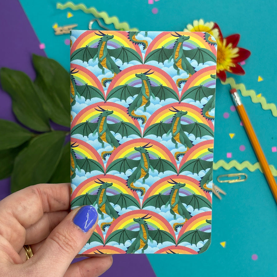 a hand holding a small notebook with rounded corners featuring a pattern of dragons, rainbows, and clouds