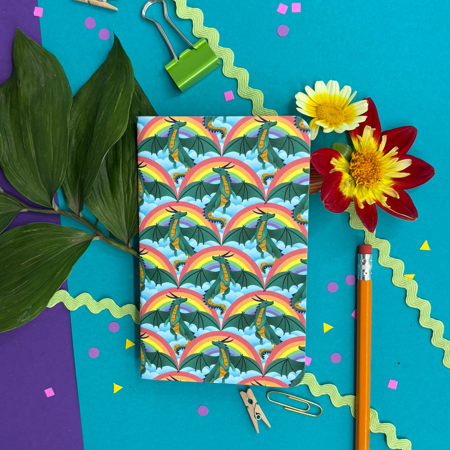 a small notebook with rounded corners featuring a pattern of dragons, rainbows, and clouds in a flat-lay on a background with flowers and stationery items