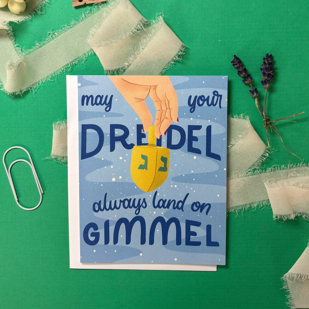 Card with a dreidel illustration and text "may your dreidel always land on gimmel" on a green background with decorative elements.