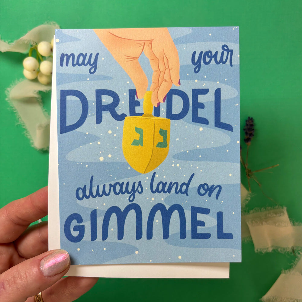 hand holding  greeting Card with a dreidel illustration and text "may your dreidel always land on gimmel" on a green background with decorative elements.