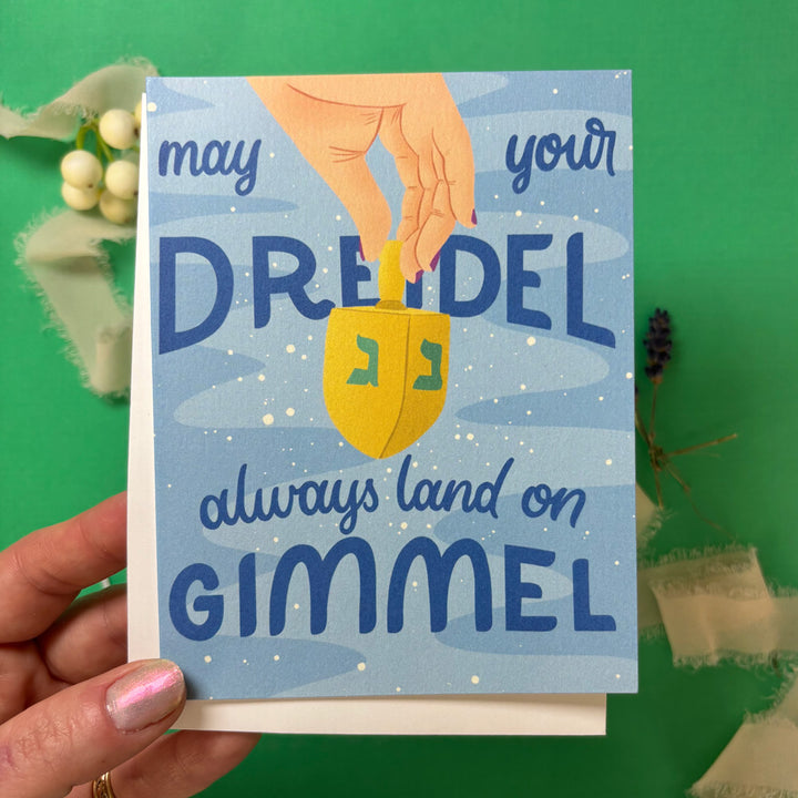 hand holding  greeting Card with a dreidel illustration and text "may your dreidel always land on gimmel" on a green background with decorative elements.