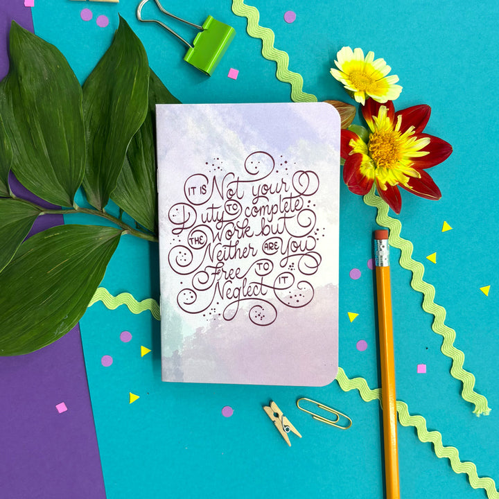 A notebook with a pastel watercolor cover and cursive quote reading 'It's not your duty to complete, nor neither you, free neglect' is styled in a flat lay on a bright teal background with green leaves, yellow and red flowers, a pencil, paper clips, and colorful confetti.