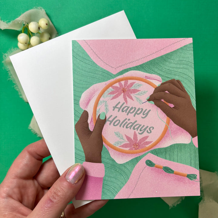 Greeting card with 'Happy Holidays' text and embroidery design, held by a hand on a green background.
