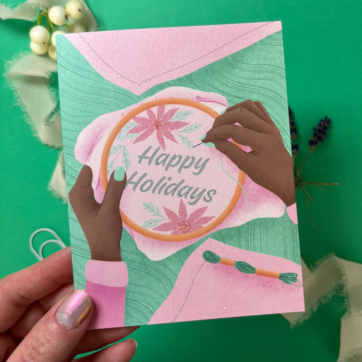 Card with embroidery design and 'Happy Holidays' text held by a hand on a green background