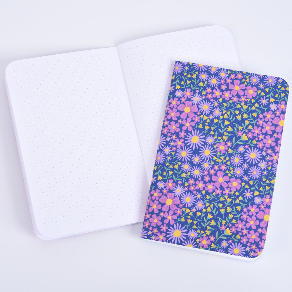 a closed small pocket notebook featuring a floral design on  dark blue background, on top of an open notebook with small dot grid pages