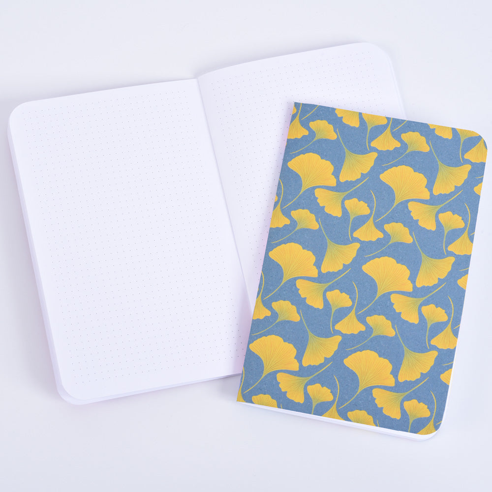 a small pocket notebook with a pattern of yellow ginkgo leaves on a blue background, resting on an open notebook with dot grid pages