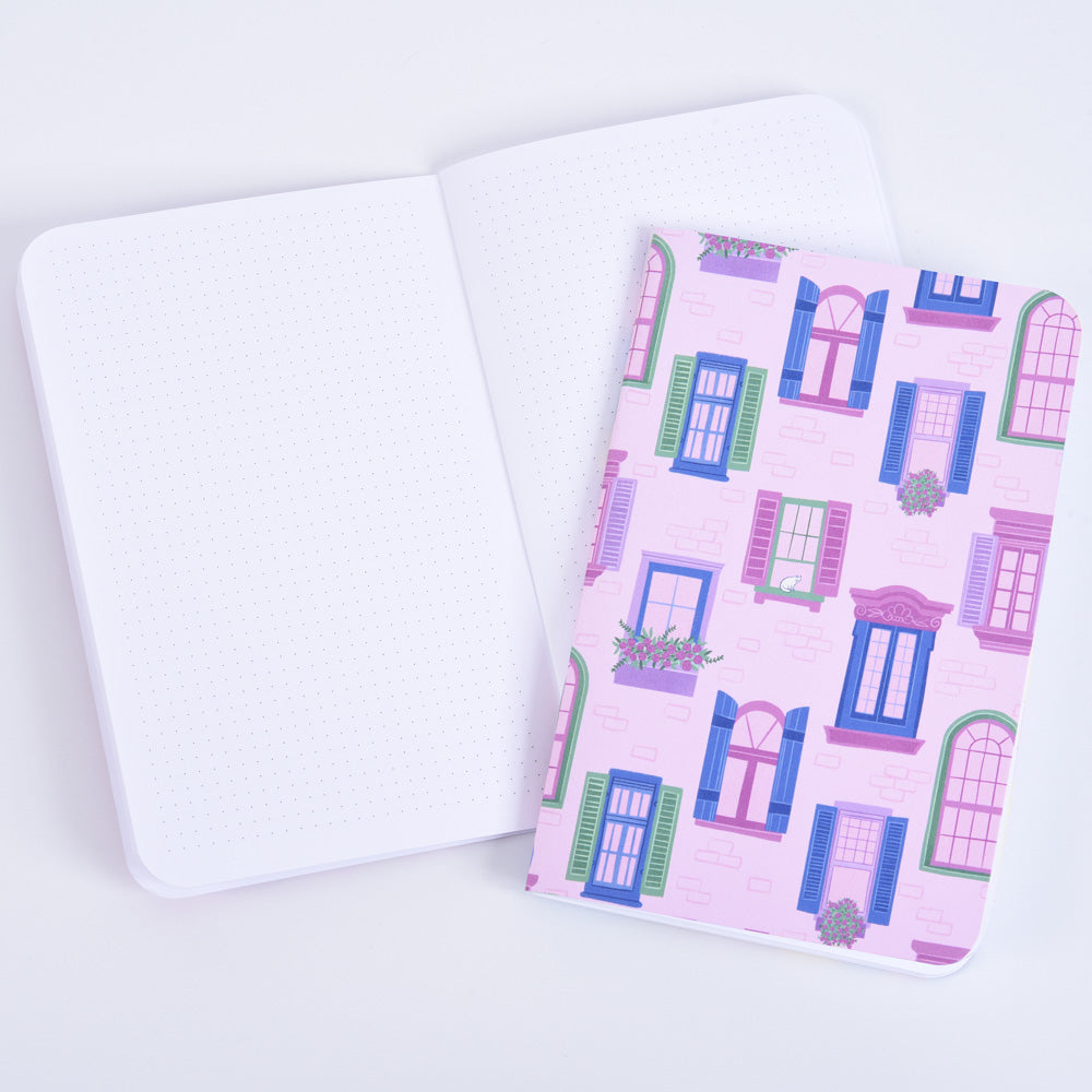 a small pocket notebook with a pattern of windows and flower boxes on a pink brick background in front of an open notebook with small dot grid paper