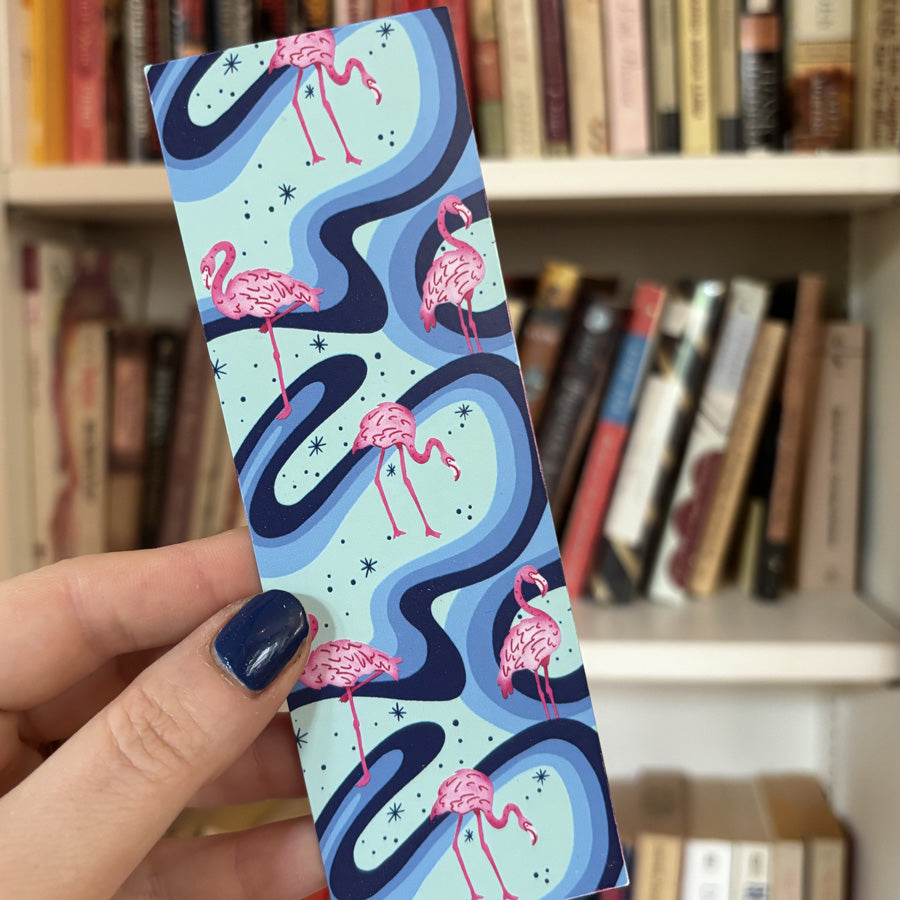 Hand holding a bookmark with flamingo design in front of a bookshelf.