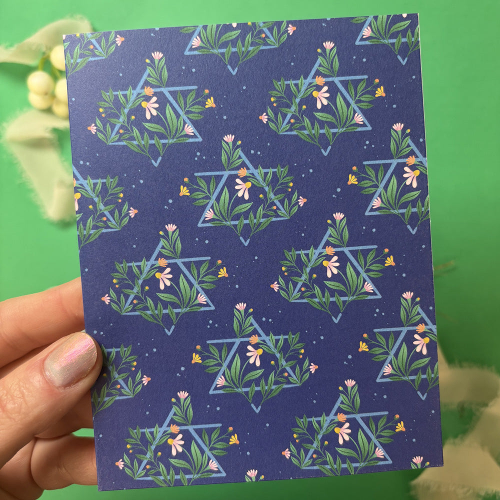 Hand holding a blue card with floral Jewish Star pattern against a green background