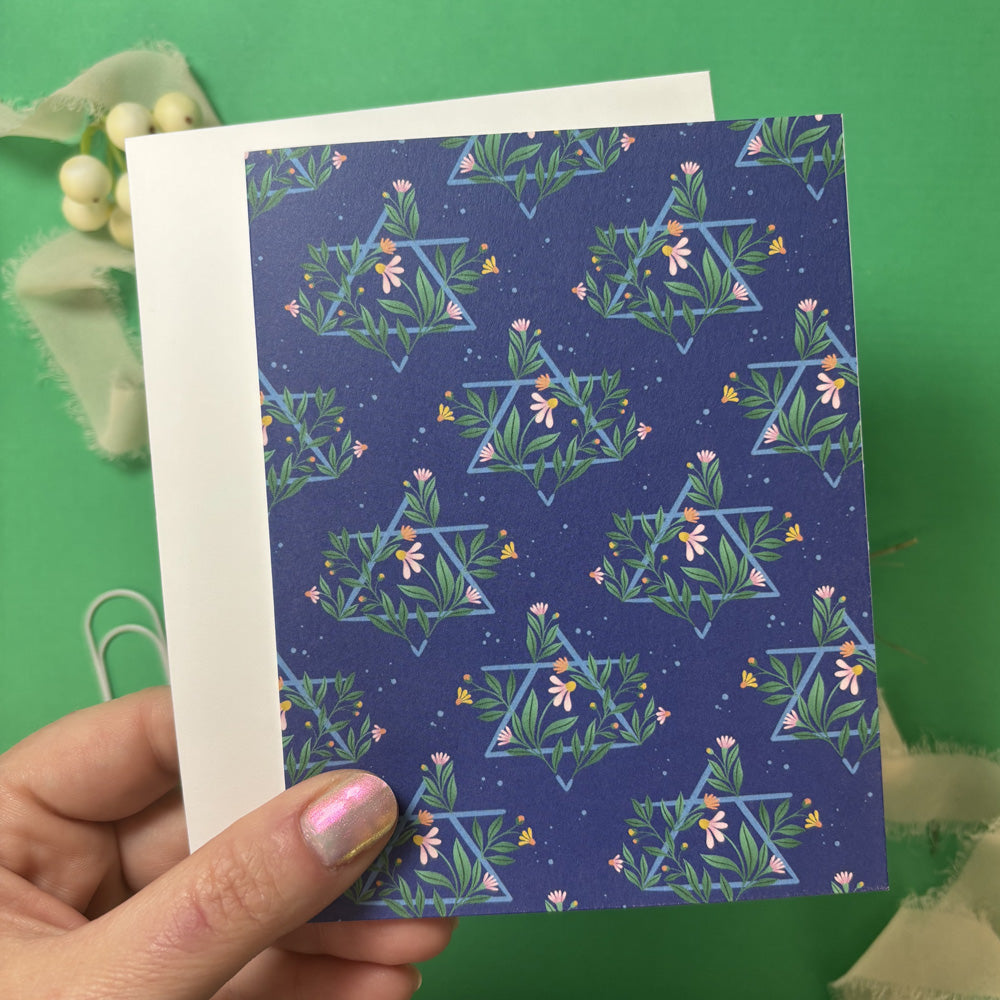 Hand holding a blue card with floral Jewish Star pattern against a green background