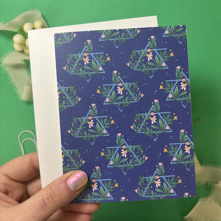Hand holding a blue card with floral Jewish Star pattern against a green background