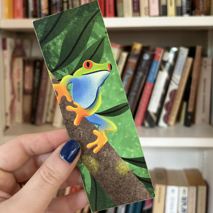Colorful frog bookmark held by a hand in front of a bookshelf.