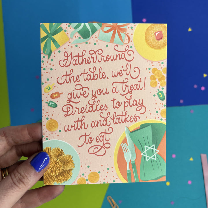 hand holding a Hanukkah card featuring illustrations items around a dinner table and script lettering "gather round the table, we'll give you a treat! Dredels to play with and latkes to eat"