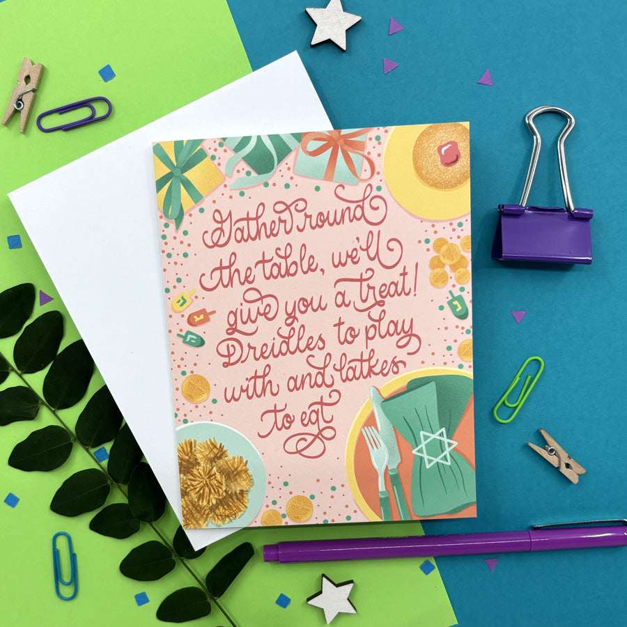 Hanukkah card featuring illustrations items around a dinner table and script lettering "gather round the table, we'll give you a treat! Dredels to play with and latkes to eat" on a colorful background with stationery items