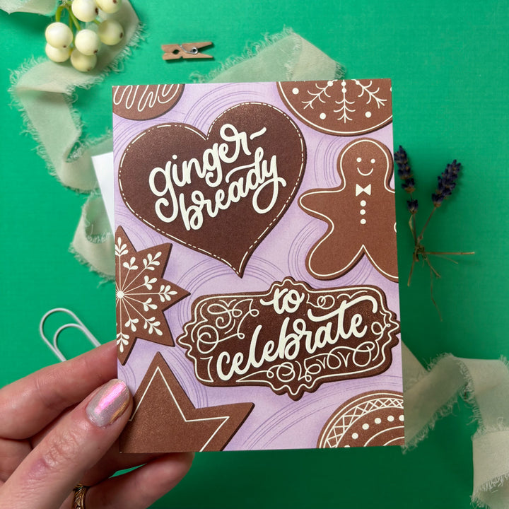 Card with gingerbread design and text "ginger-bready to celebrate" held by a hand on a green background