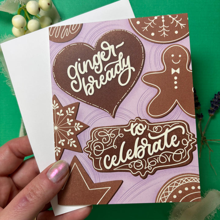 Greeting card with gingerbread design and text 'Ginger Bready to Celebrate' held by a hand on a green background.
