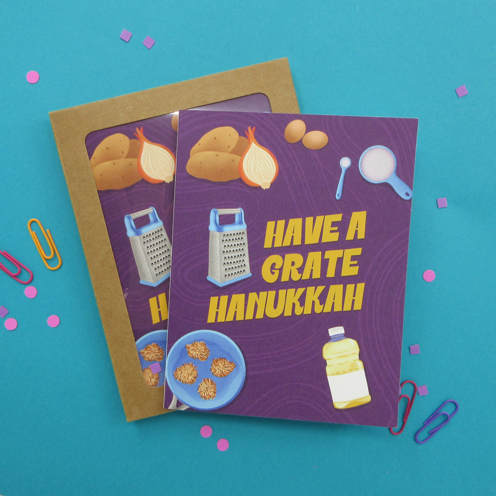 a box set of greeting cards with a Hanukkah theme are arranged on a bright blue background with colorful paper clips and confetti. The front card has a purple background with yellow text reading 'HAVE A GRATE HANUKKAH' and illustrations of food and kitchen items.