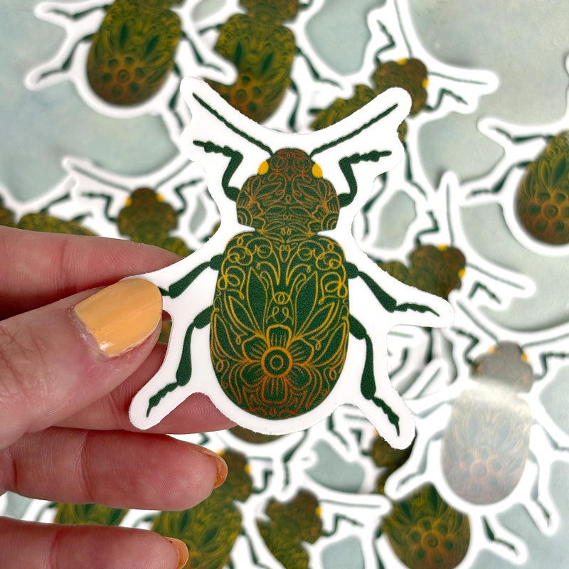 hand holding A sticker depicting a green beetle with floral-like patterns on its body.